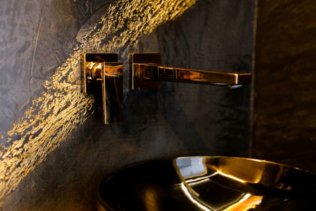 Kreative Wandgestaltung an einer mit dem Kalkputz Terra Nuovo verputzten Badezimmerwand: Die Wand ist in Anthrazit gestaltet und hat einen goldenen Akzent, der wirkt wie aufgewischt. An der gleichen Stelle hängen an der Wand ein goldener Wasserhahn sowie der zugehörige Griff. Ein goldenes Waschbecken ist im Anschnitt zu sehen.