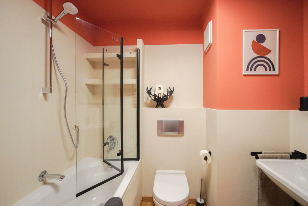 Das Nachher-Bild zeigt das gleiche Badezimmer mit fugenlosen, wasserabweisenden Wänden aus HardRock Pro.  Die Hauptfarbe ist Creme mir einem einem Absatz in Pfirsich-Orange. Neben den neuen Wänden sind auch neue, moderne Sanitärprodukten und ein farblich passendes Wandbild zu sehen.